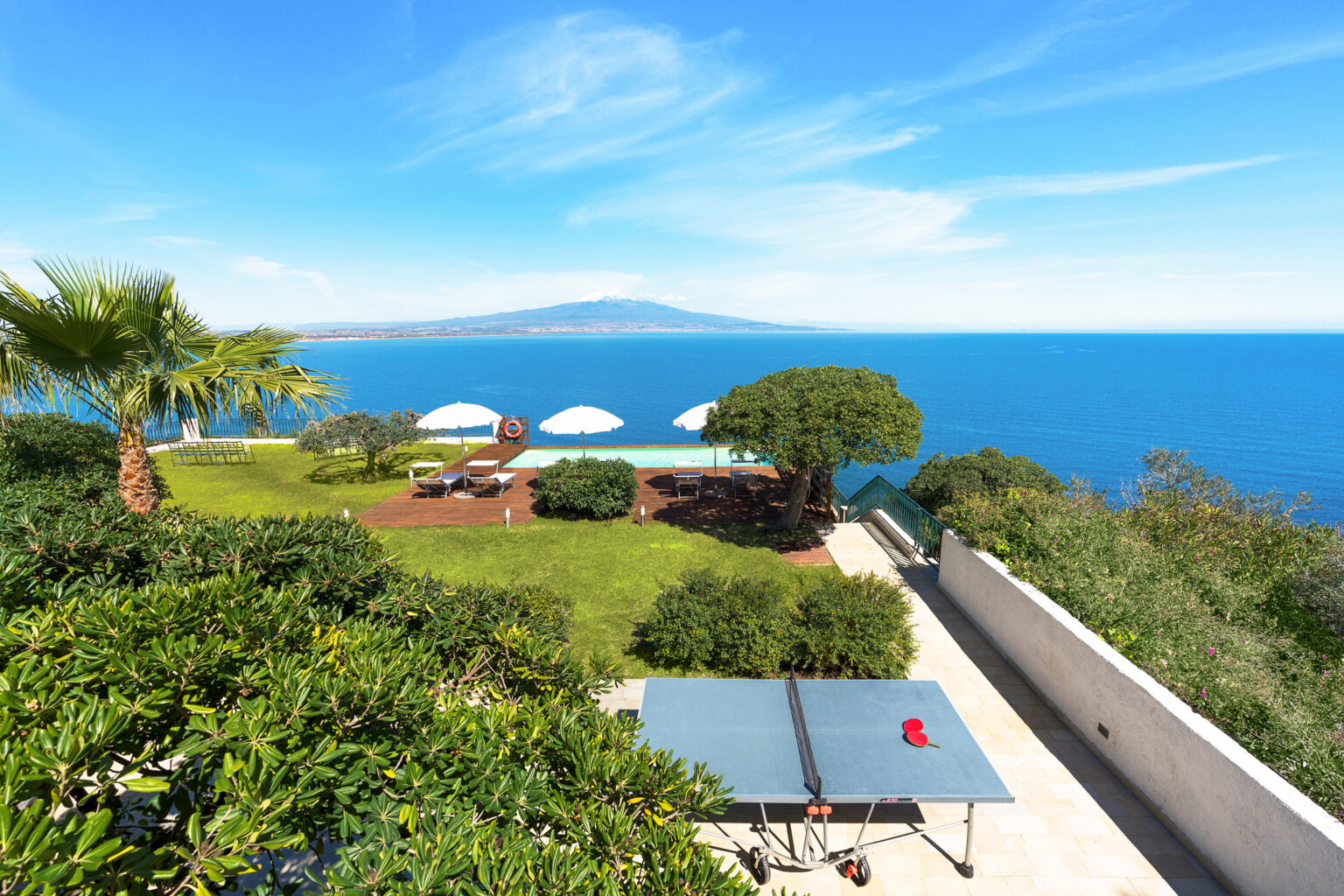 Scenic view of a coastal villa with a spacious garden, featuring a ping pong table, sun loungers, and umbrellas overlooking the blue sea and distant mountains. | Only on SopranoVillas