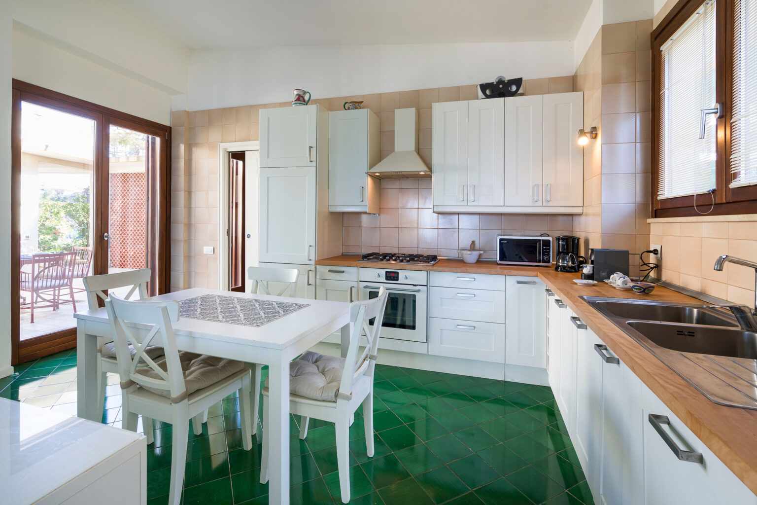 Bright and modern kitchen featuring white cabinetry, a wooden countertop, and a dining table with white chairs. The space is illuminated by natural light from large windows, complemented by green flooring. Ideal for cooking and entertaining. | Only on SopranoVillas