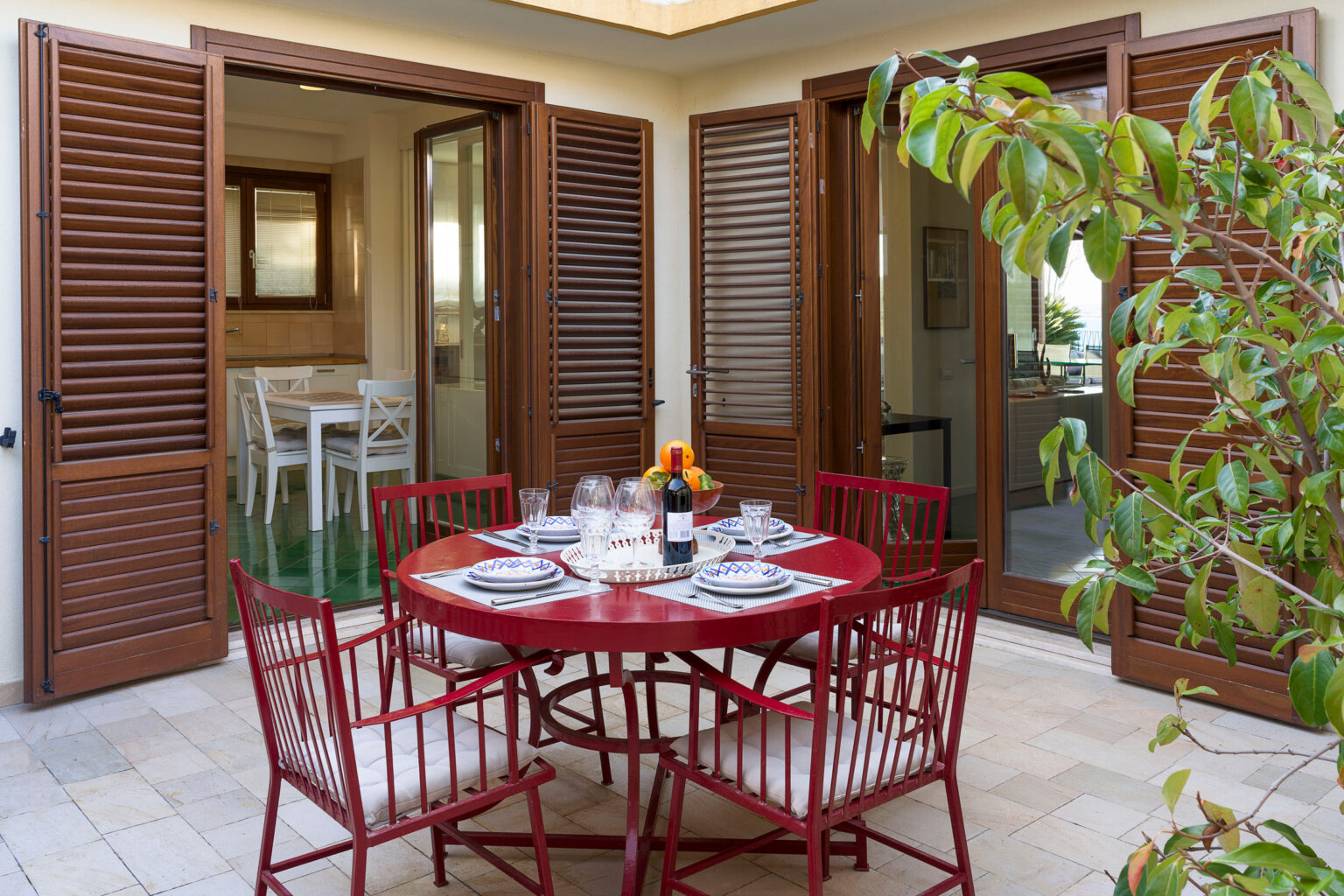 Outdoor dining area featuring a round red table set for a meal, surrounded by matching chairs. The space is framed by wooden shutters and includes lush greenery, creating a welcoming atmosphere for al fresco dining. | Only on SopranoVillas