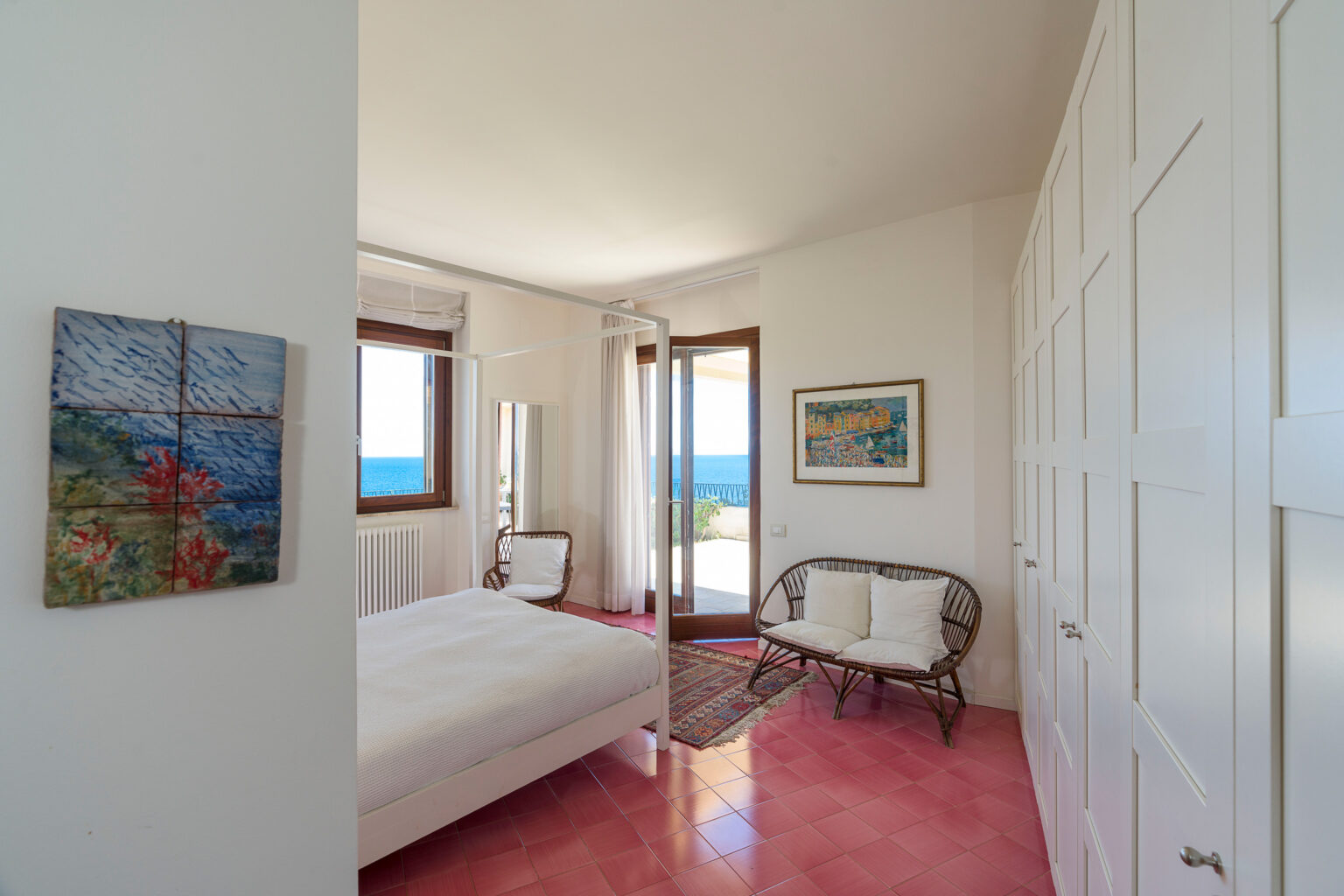 Cozy bedroom with a white bed, wicker chair, and large windows showcasing a scenic ocean view, featuring light-colored walls and pink flooring, complemented by decorative artwork. | Only on SopranoVillas
