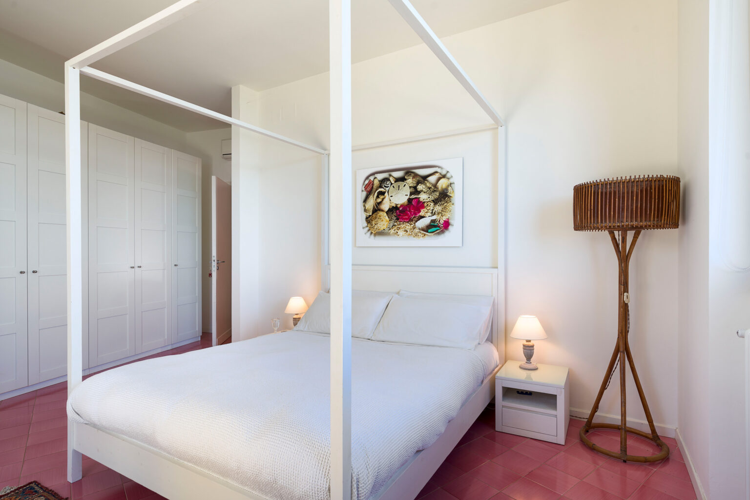Modern bedroom featuring a white four-poster bed, decorative artwork on the wall, a wooden floor lamp, and a nightstand with a lamp, complemented by light-colored walls and pink carpeting. | Only on SopranoVillas