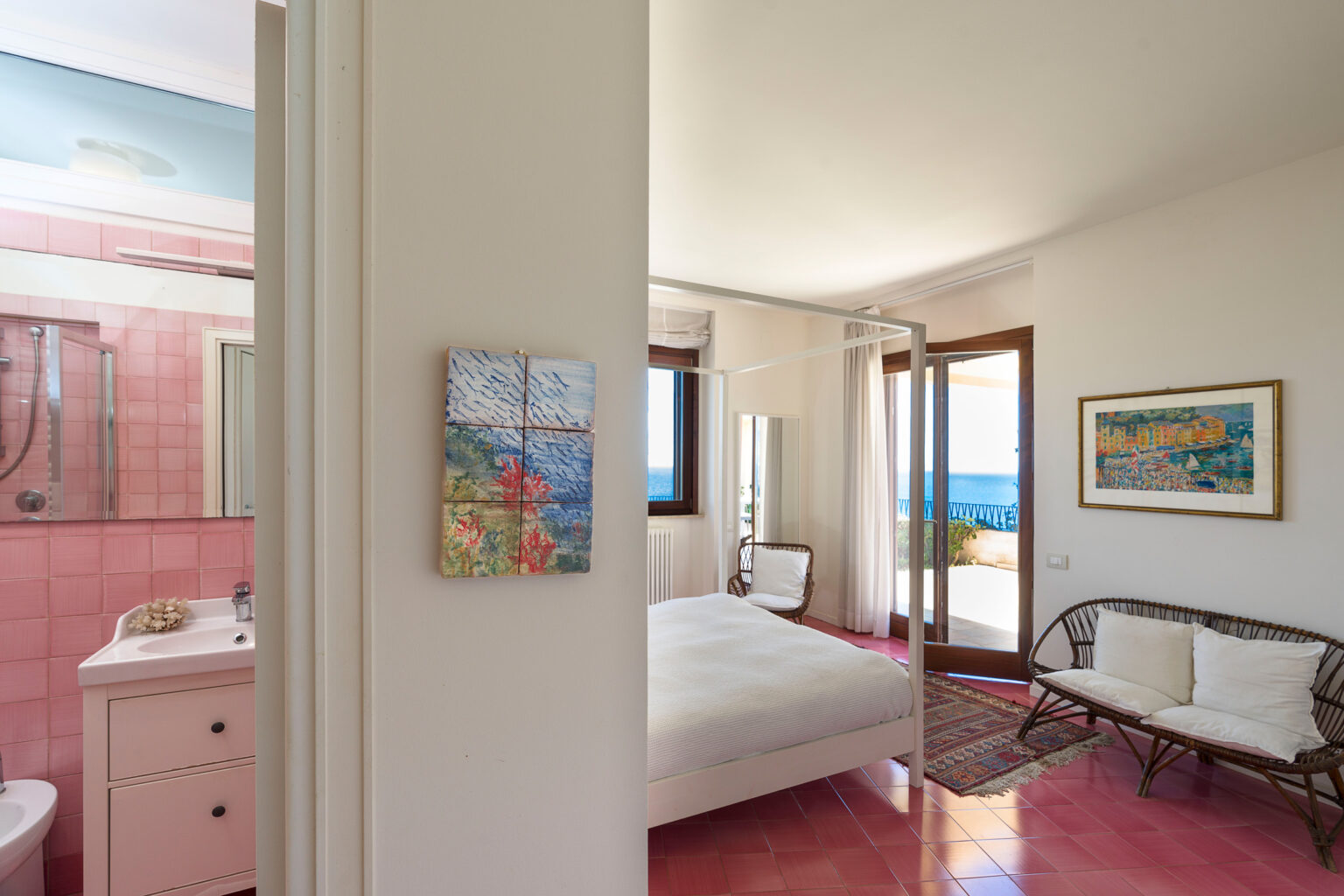 Cozy bedroom with a four-poster bed, featuring a view of the sea through large windows. The room includes a comfortable seating area with wicker chairs, a colorful area rug, and a bathroom visible in the background with pink tile accents. Decorative artwork adorns the walls, enhancing the inviting atmosphere. | Only on SopranoVillas