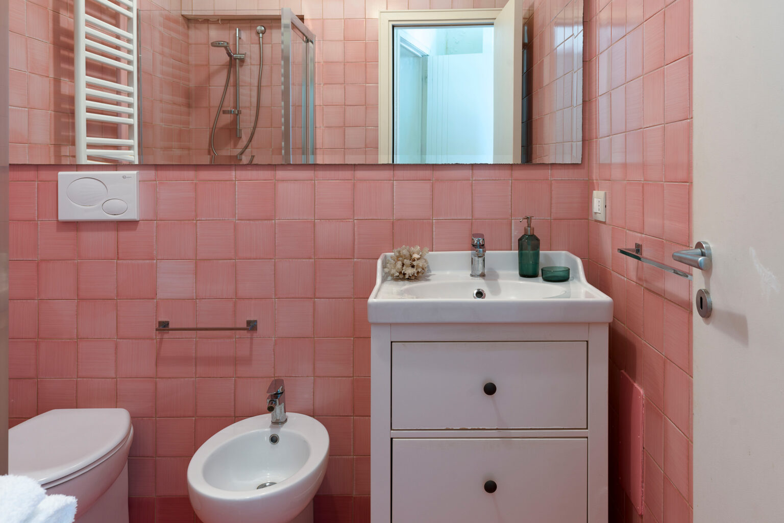 Small modern bathroom featuring pink tiled walls, a white sink with drawers, a bidet, and a shower area with glass door. Natural light enters through a mirror, enhancing the space's brightness. | Only on SopranoVillas
