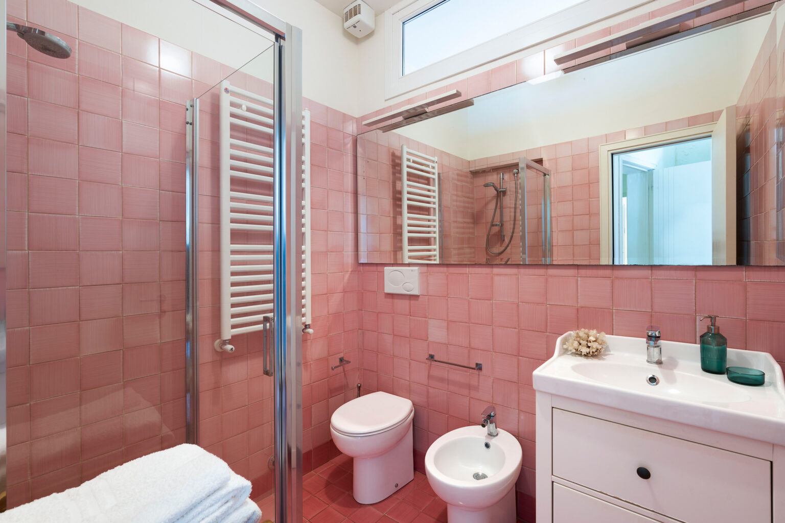 Modern bathroom featuring pink tiled walls, a glass shower enclosure, a white vanity with storage, and a bidet. Soft white towels are neatly arranged, enhancing the clean and inviting atmosphere. | Only on SopranoVillas