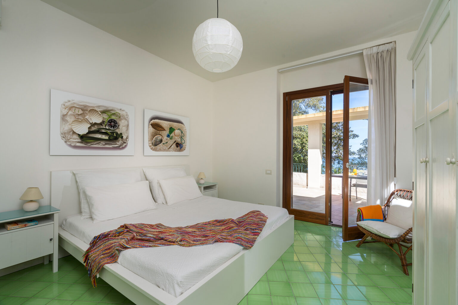 Bright and airy bedroom featuring a white bed with plush pillows and a colorful throw, complemented by coastal-themed artwork on the walls. The room boasts green tiled flooring and large glass doors that open to a sunny outdoor patio, with natural light streaming in. Ideal for a relaxing getaway. | Only on SopranoVillas