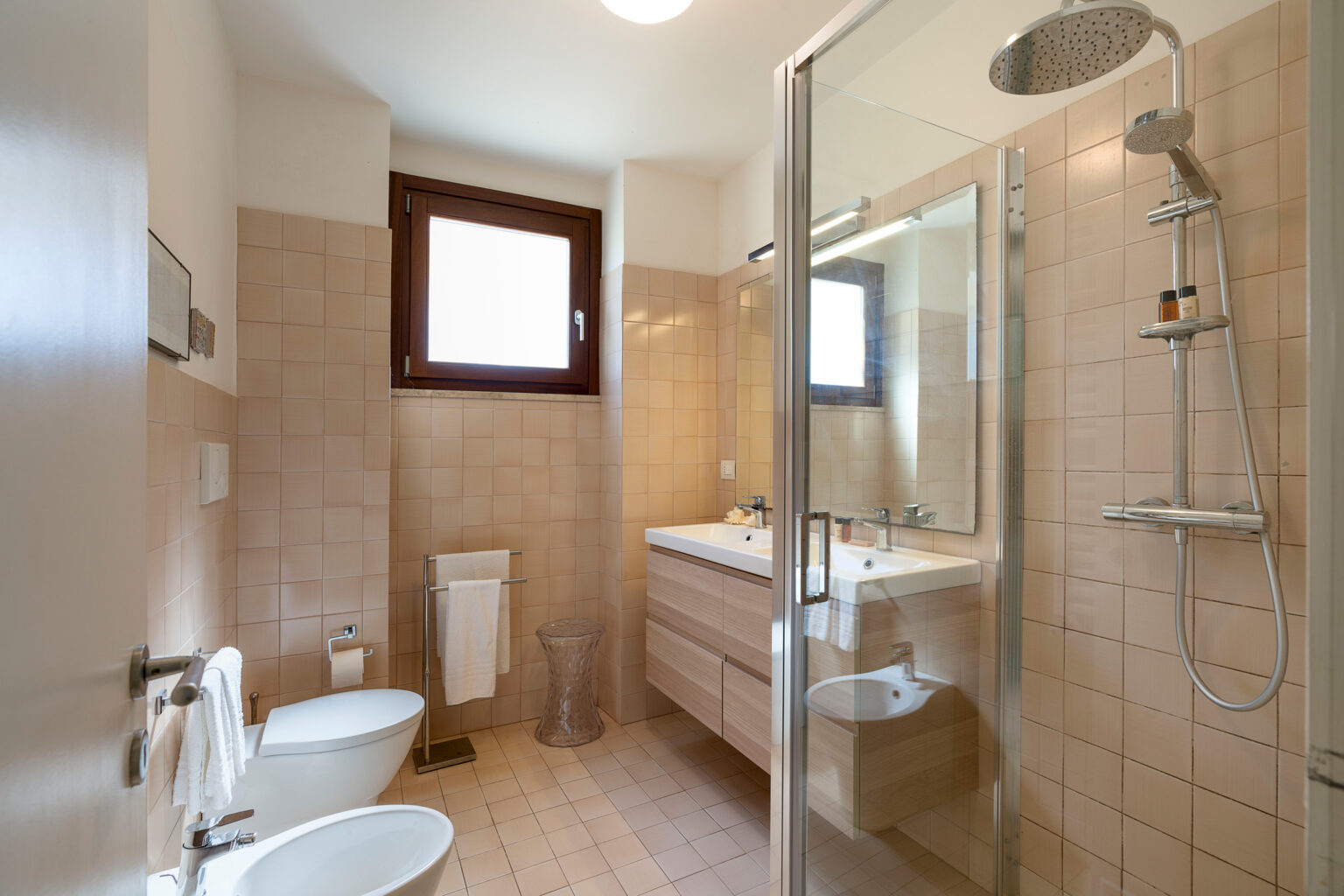 Modern bathroom featuring a glass shower enclosure, double sink vanity with wooden cabinetry, beige tiled walls, and natural light streaming through a window. The space includes a bidet and towel rack, creating a clean and functional design. | Only on SopranoVillas