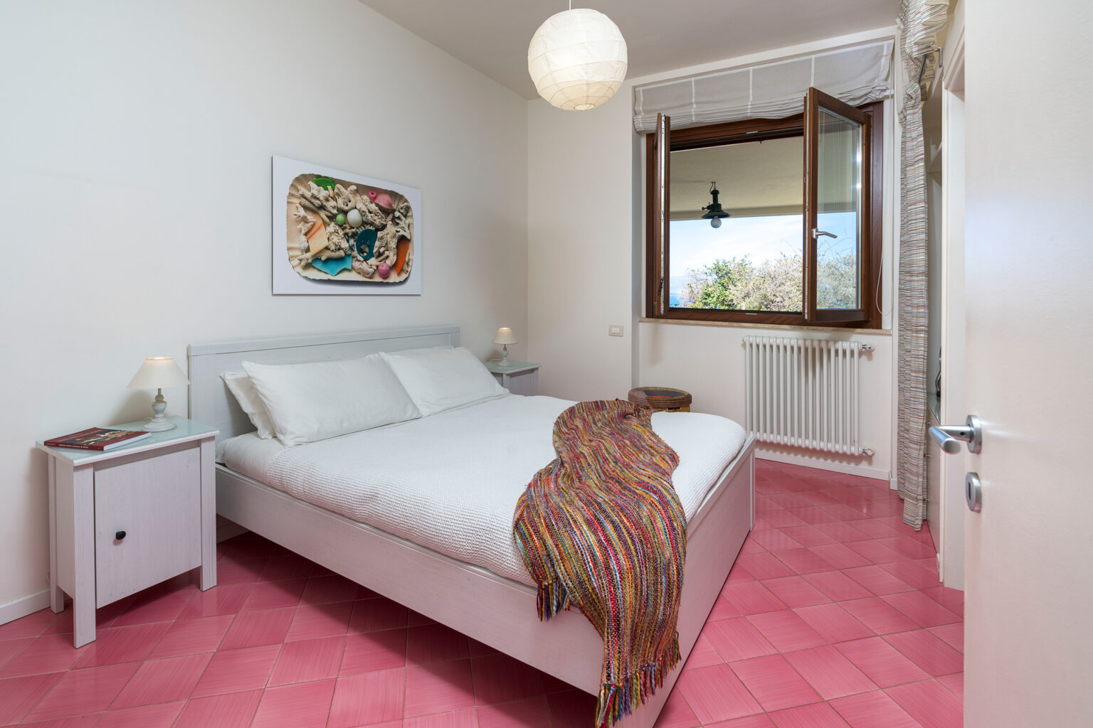 Modern bedroom featuring a double bed with white linens, a colorful throw blanket, pink tiled flooring, and a large window with natural light. Decorated with artwork on the wall and a bedside table with a lamp, this inviting space emphasizes comfort and style. | Only on SopranoVillas