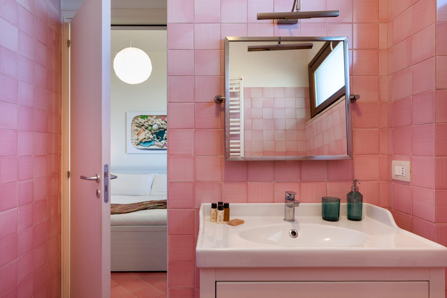 Modern pink-tiled bathroom featuring a white sink, mirror, and decorative bottles, with a glimpse of a bedroom visible through an open door. | Only on SopranoVillas