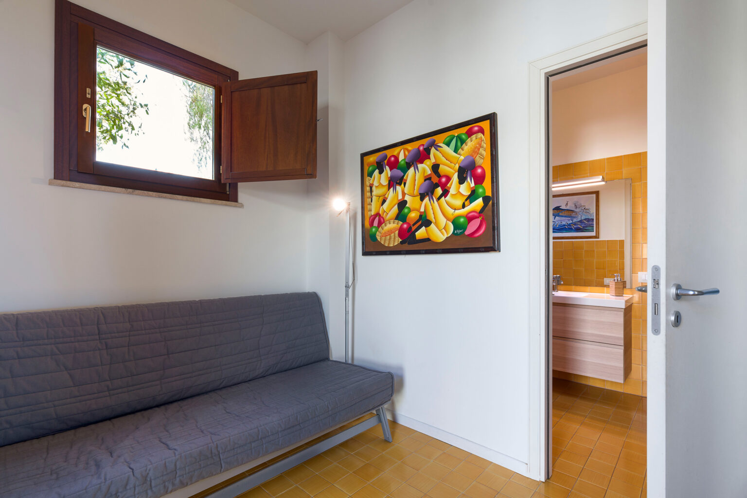Cozy interior view of a room featuring a gray sofa, vibrant fruit-themed artwork on the wall, and a doorway leading to a bathroom. Natural light enters through an open window, enhancing the inviting atmosphere. | Only on SopranoVillas