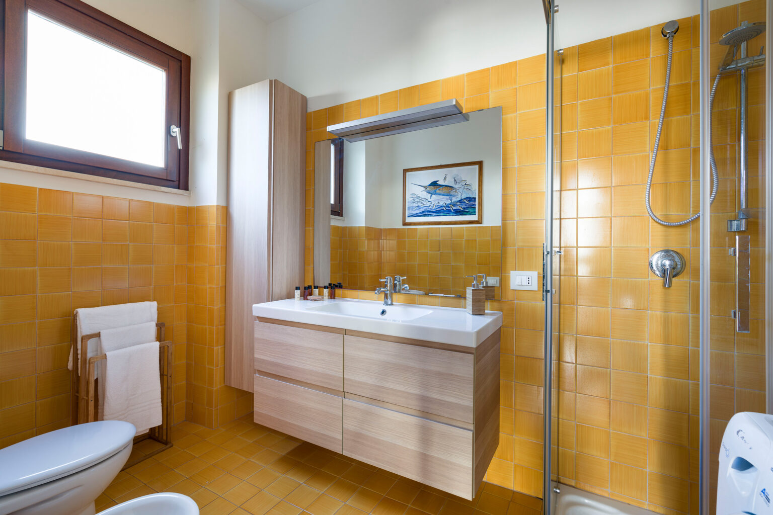 Modern bathroom featuring a wooden vanity with double sinks, a glass shower enclosure, and bright yellow tiled walls. Natural light streams in through a window, enhancing the spacious feel of the room. | Only on SopranoVillas