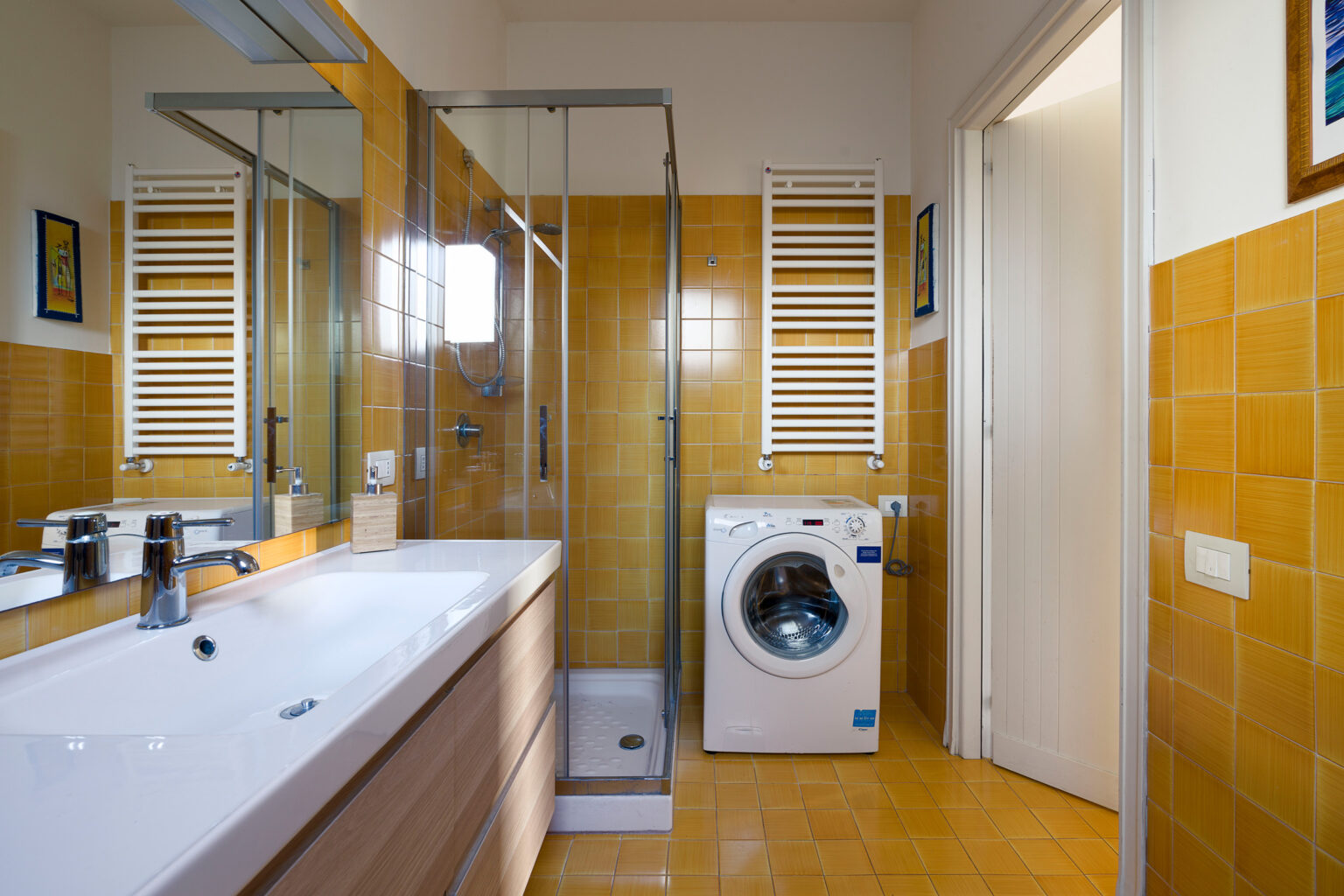 Modern bathroom featuring a glass shower enclosure, a sleek vanity with a sink, a washing machine, and warm yellow tile accents. The design emphasizes functionality and contemporary style. | Only on SopranoVillas