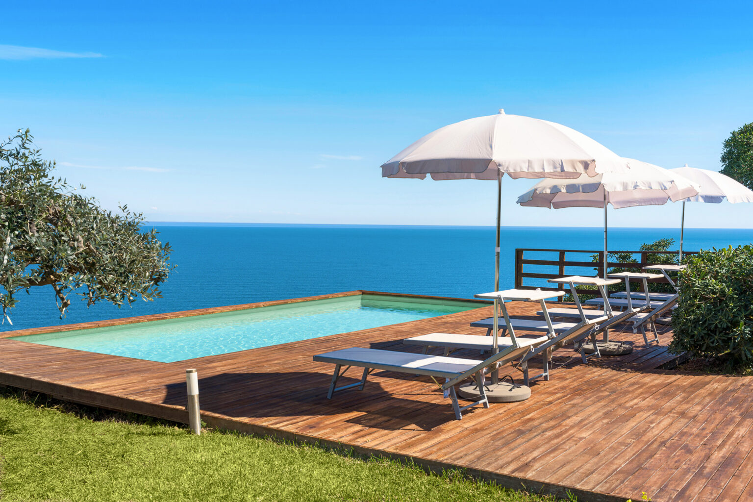 Luxury poolside view overlooking the ocean, featuring sun loungers and umbrellas on a wooden deck, surrounded by lush greenery and a clear blue sky. | Only on SopranoVillas