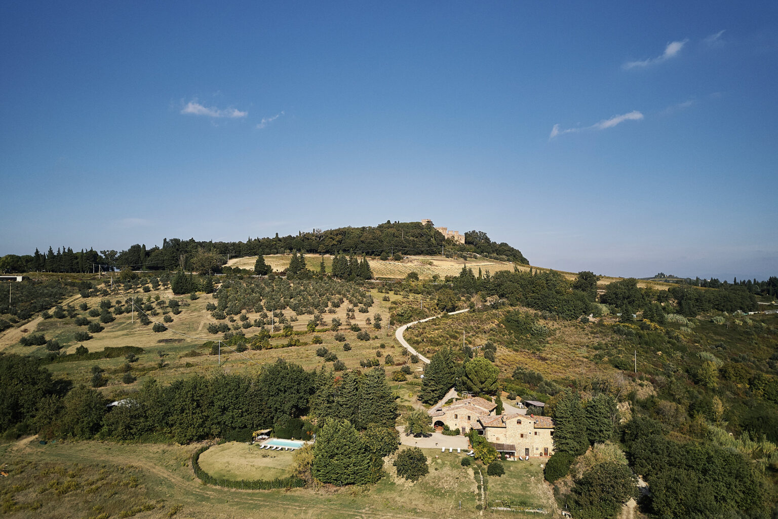 Aerial view of a scenic Tuscan landscape featuring rolling hills, olive groves, and a rustic stone villa with a swimming pool, set against a clear blue sky. The image captures the natural beauty and tranquility of the Italian countryside. | Only on SopranoVillas