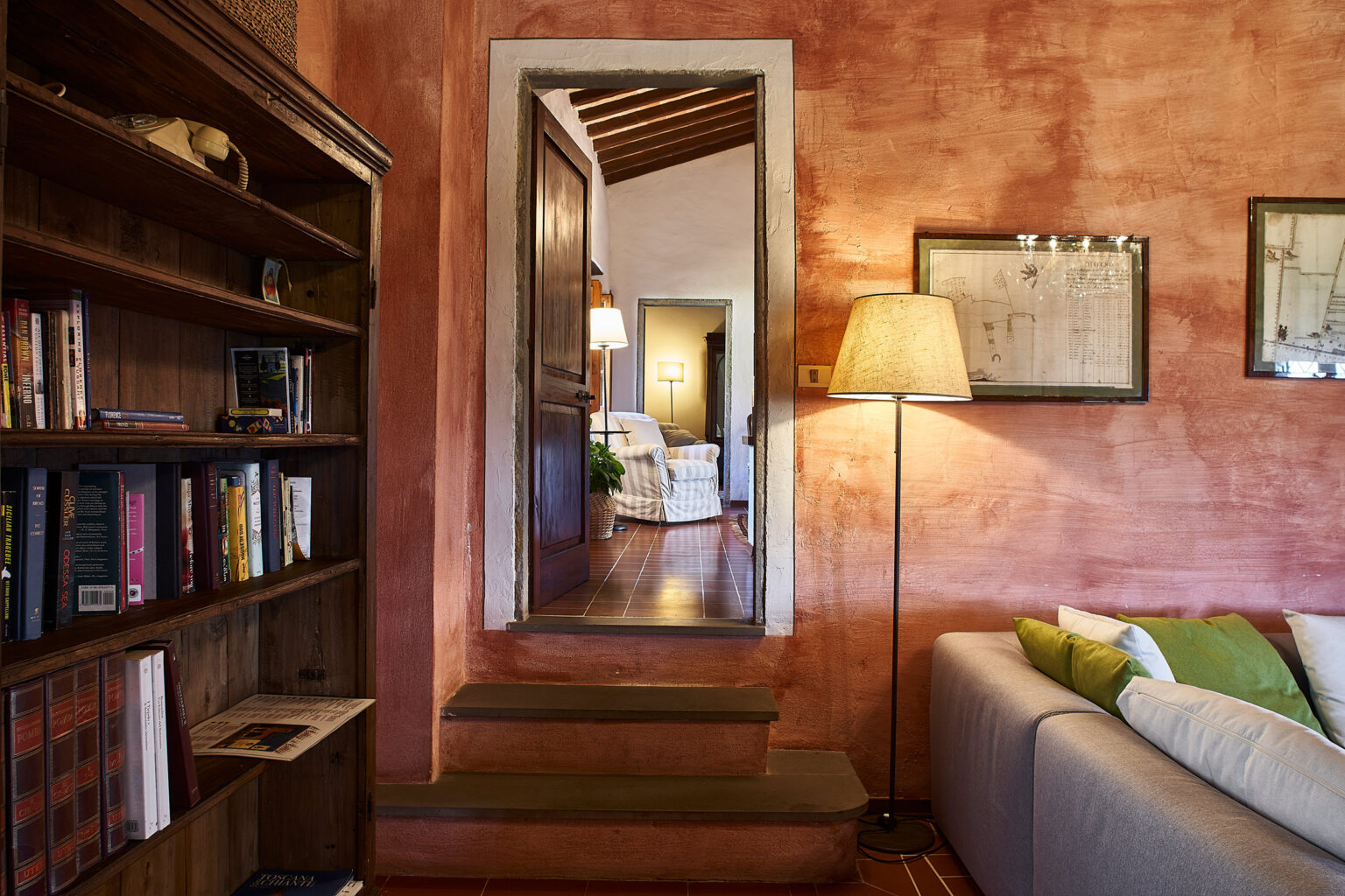 Cozy living room interior featuring a wooden bookshelf filled with books, a warm floor lamp, and a view of a doorway leading to another room with comfortable seating. The walls are painted in a rich, textured red, enhancing the inviting atmosphere. | Only on SopranoVillas
