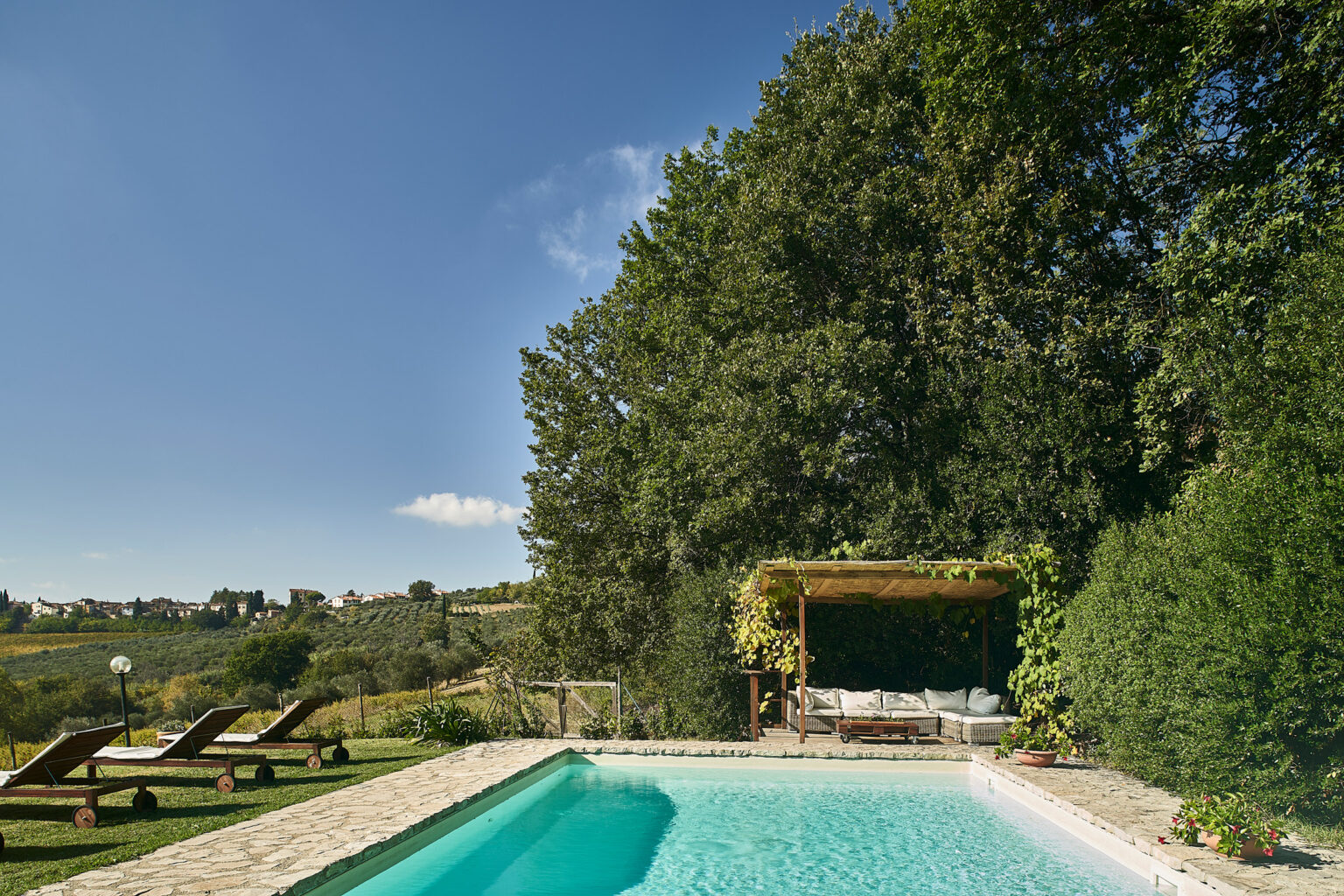 Serene outdoor pool area surrounded by lush greenery, featuring comfortable lounge chairs and a shaded seating area under a pergola, ideal for relaxation and enjoying nature. | Only on SopranoVillas
