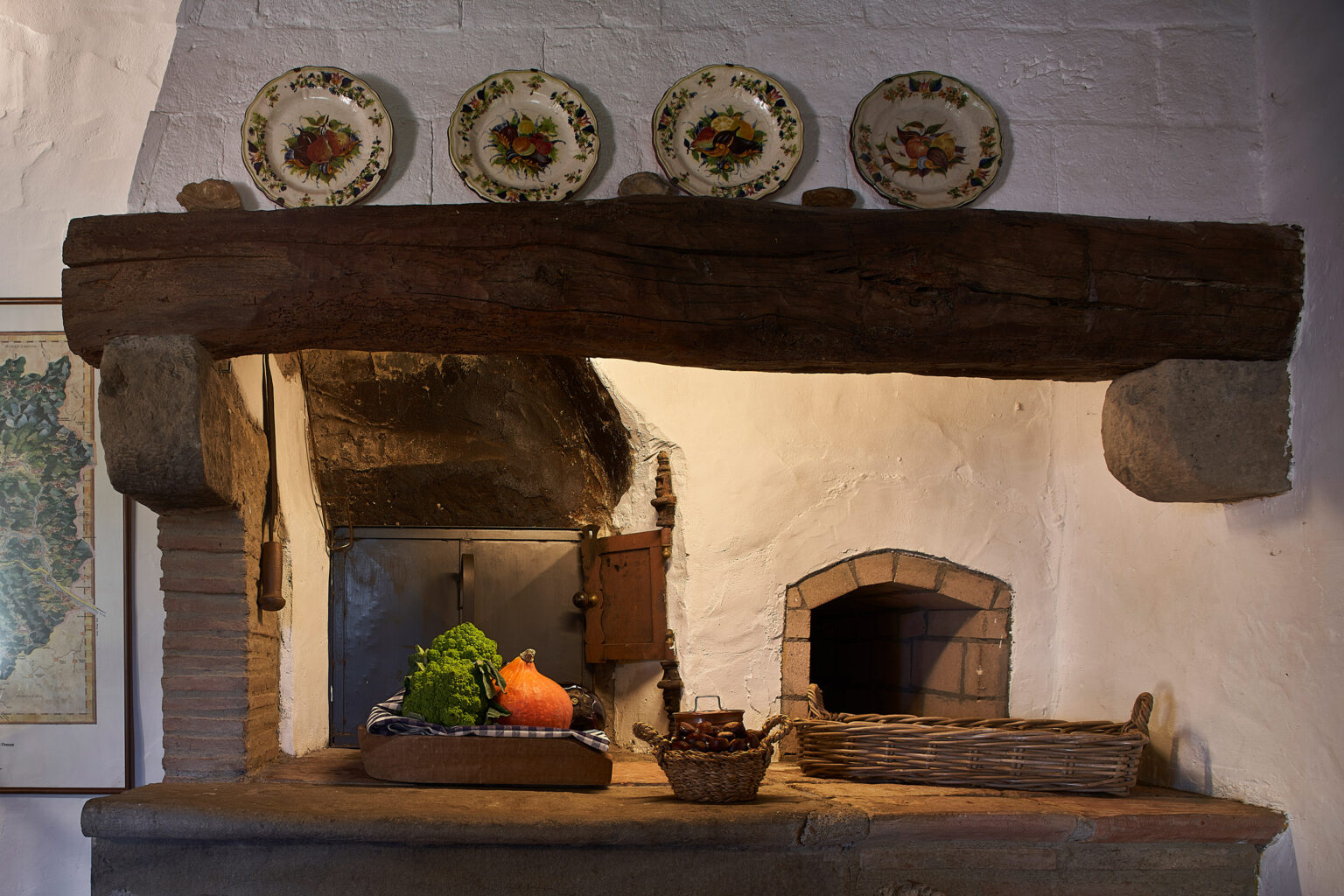 Rustic kitchen interior featuring a wooden beam with decorative plates, a stone countertop displaying fresh vegetables including broccoli and pumpkin, and woven baskets. The background includes a vintage map and a traditional oven, highlighting a cozy, inviting atmosphere. | Only on SopranoVillas
