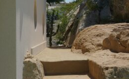 Narrow pathway leading between a white wall and a rocky terrain, surrounded by greenery, ideal for nature walks or outdoor exploration.