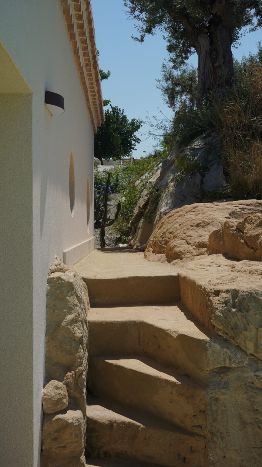 Narrow pathway leading between a white wall and a rocky terrain, surrounded by greenery, ideal for nature walks or outdoor exploration. | Only on SopranoVillas