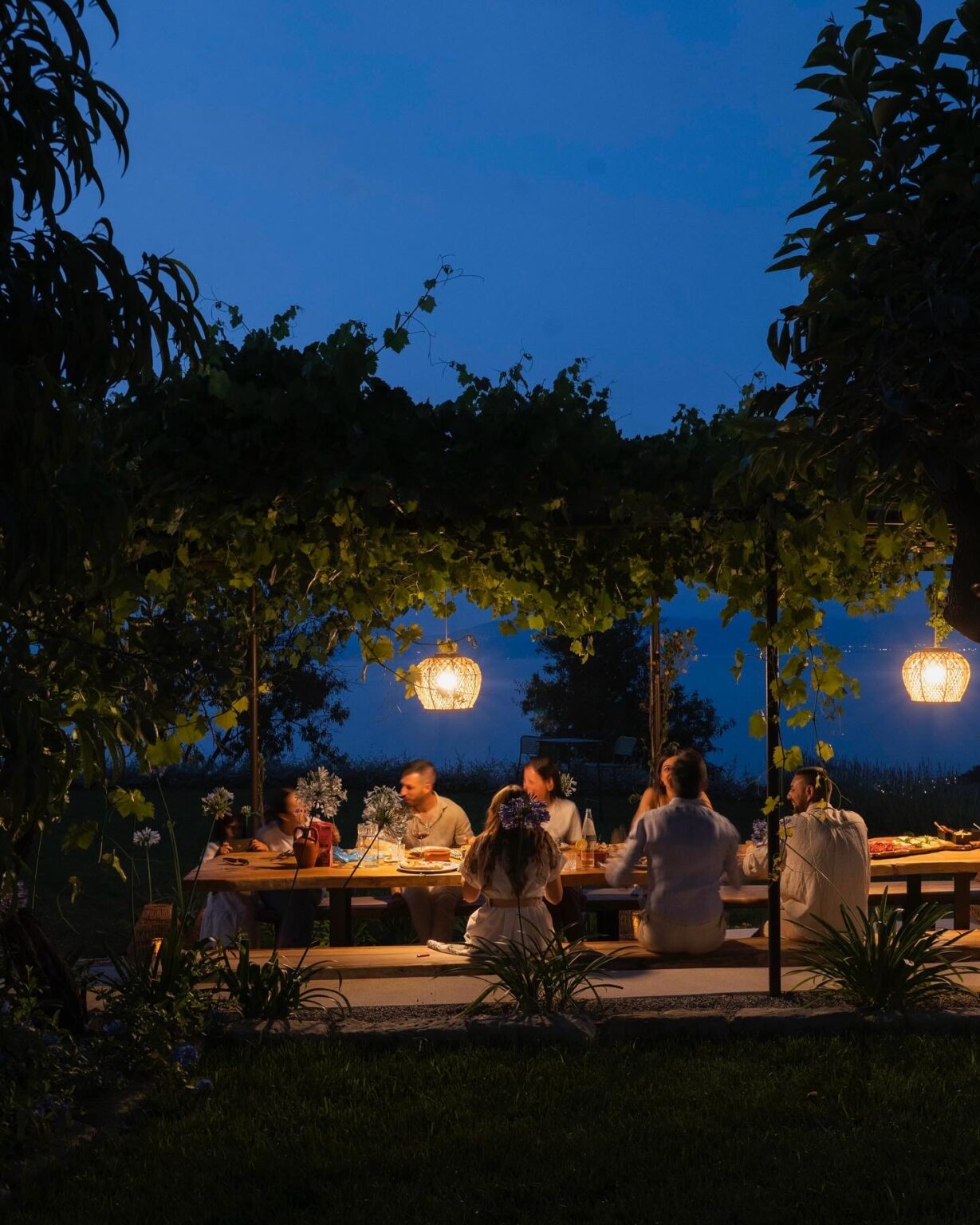 Group of people enjoying a dinner gathering under a vine-covered pergola at night, illuminated by hanging lanterns, creating a warm and inviting atmosphere. | Only on SopranoVillas