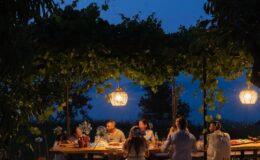 Group of people enjoying a dinner gathering under a vine-covered pergola at night, illuminated by hanging lanterns, creating a warm and inviting atmosphere.