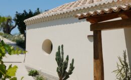 Modern white building with round window, surrounded by cacti and greenery in a sunny outdoor setting.