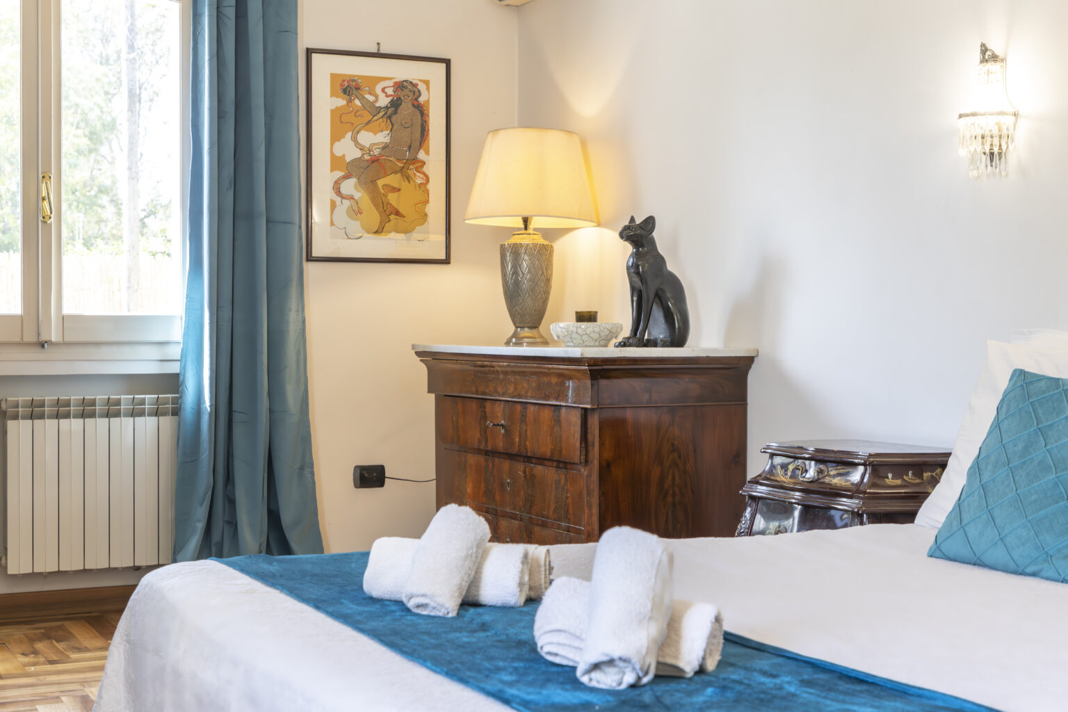 Cozy bedroom featuring a white bed with turquoise accents, neatly rolled towels, a wooden nightstand with a decorative cat figurine, and a warm lamp, complemented by elegant curtains and artwork on the wall. | Only on SopranoVillas