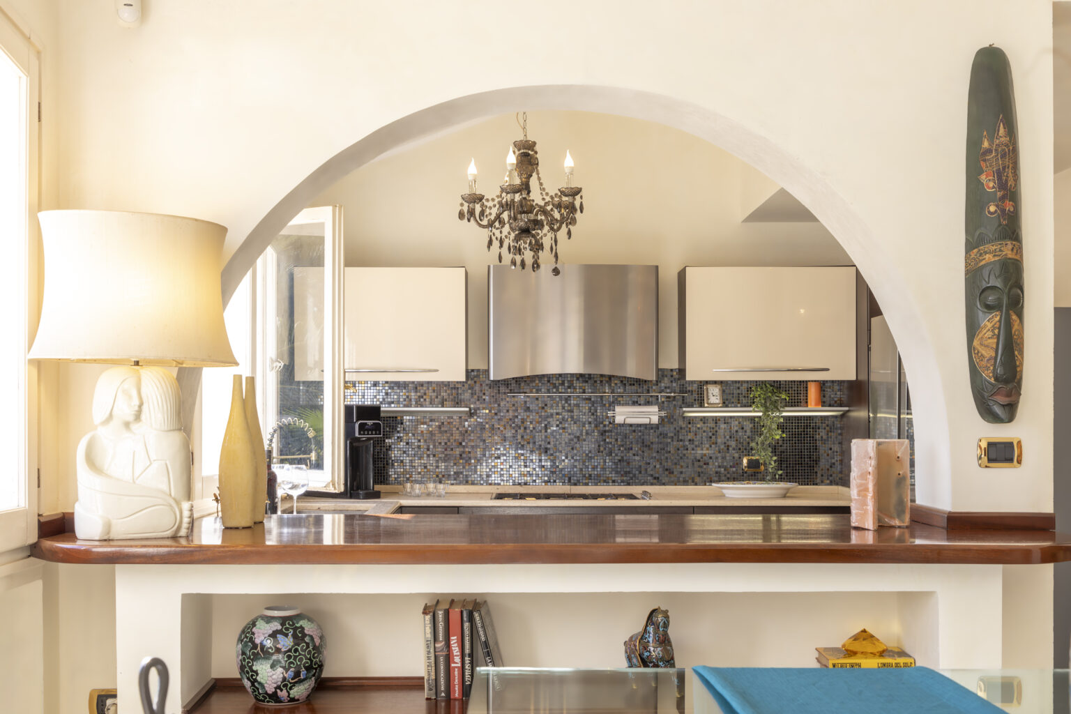 Modern kitchen interior featuring a stylish archway, elegant chandelier, and contemporary appliances. Decorative lamp and artwork add a touch of personality to the space. | Only on SopranoVillas