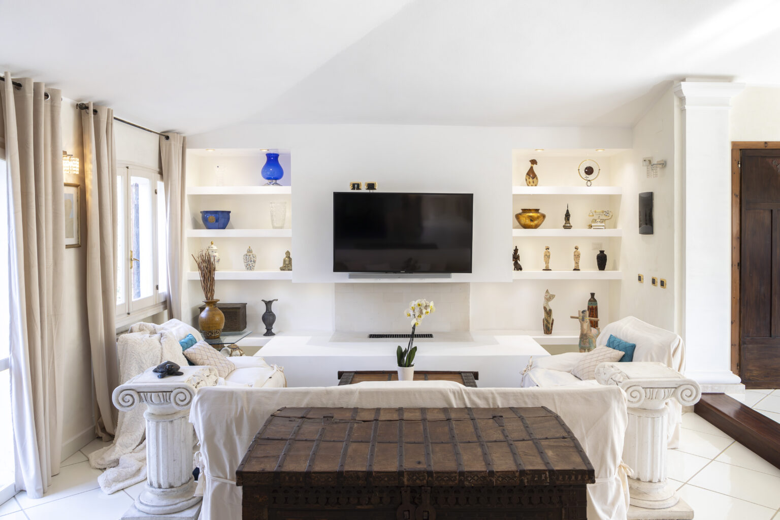 Modern living room with white walls, stylish shelving displaying decorative items, a flat-screen TV, and comfortable seating. Natural light streams in through large windows, creating a bright and inviting atmosphere. | Only on SopranoVillas