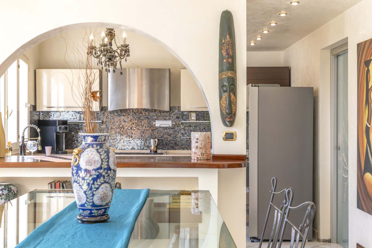 Bright and modern kitchen interior featuring a blue and white decorative vase on a glass dining table, stainless steel appliances, and an artistic wall decor. The space is illuminated with natural light and highlights a stylish design with an arched entryway. | Only on SopranoVillas