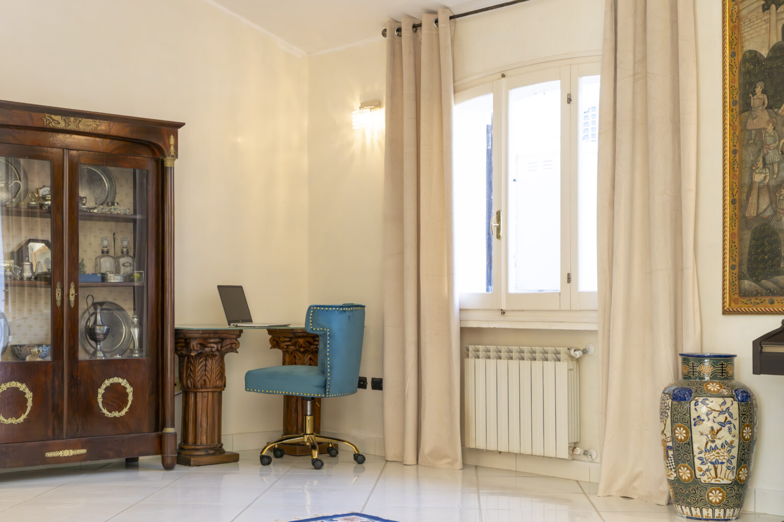 Elegant home office setup featuring a blue chair at a wooden desk, a glass display cabinet, and large windows with light curtains, creating a bright and inviting workspace. | Only on SopranoVillas