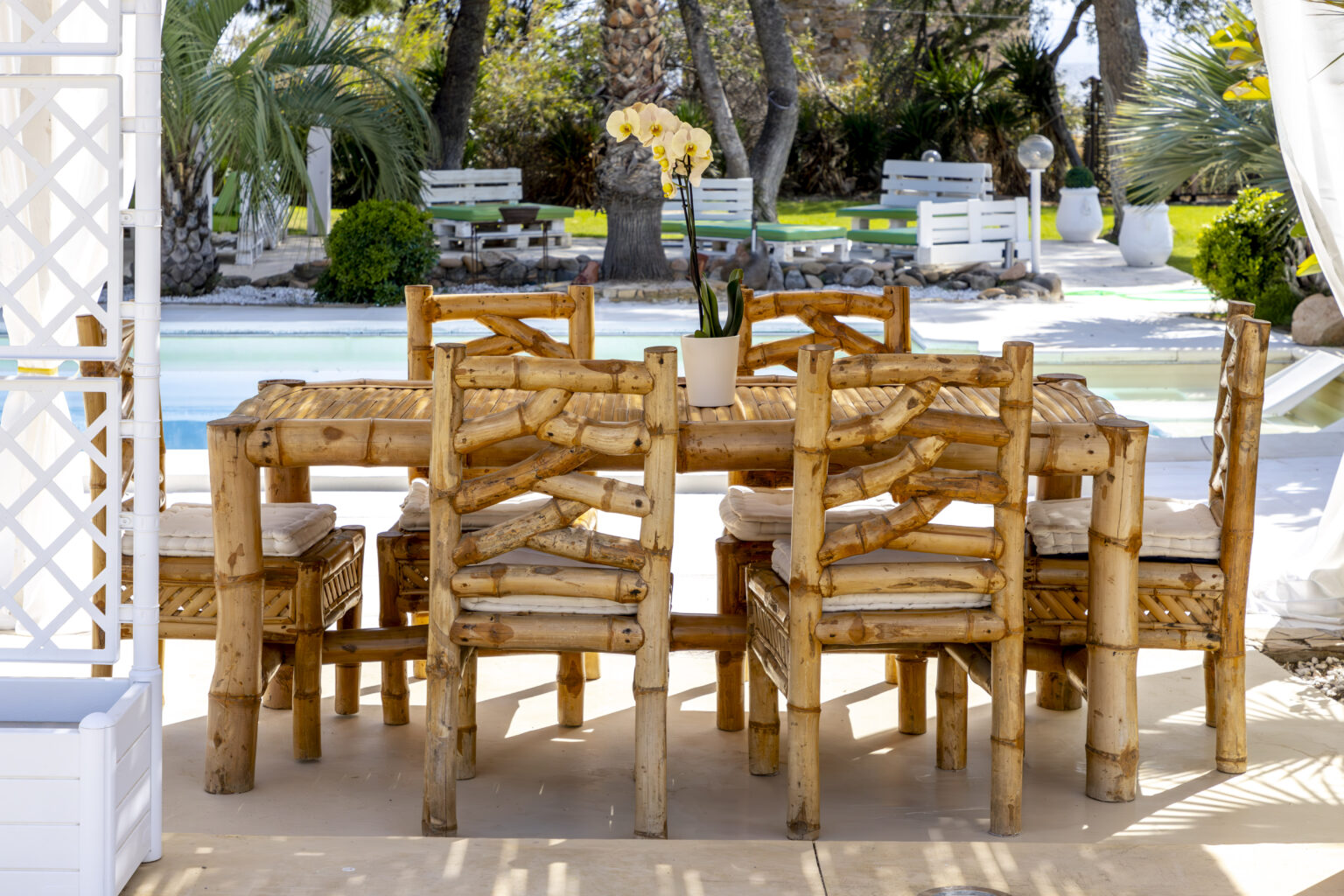 Bamboo dining table set with eight chairs, surrounded by a serene outdoor setting featuring a swimming pool and lush greenery. Perfect for alfresco dining and gatherings. | Only on SopranoVillas