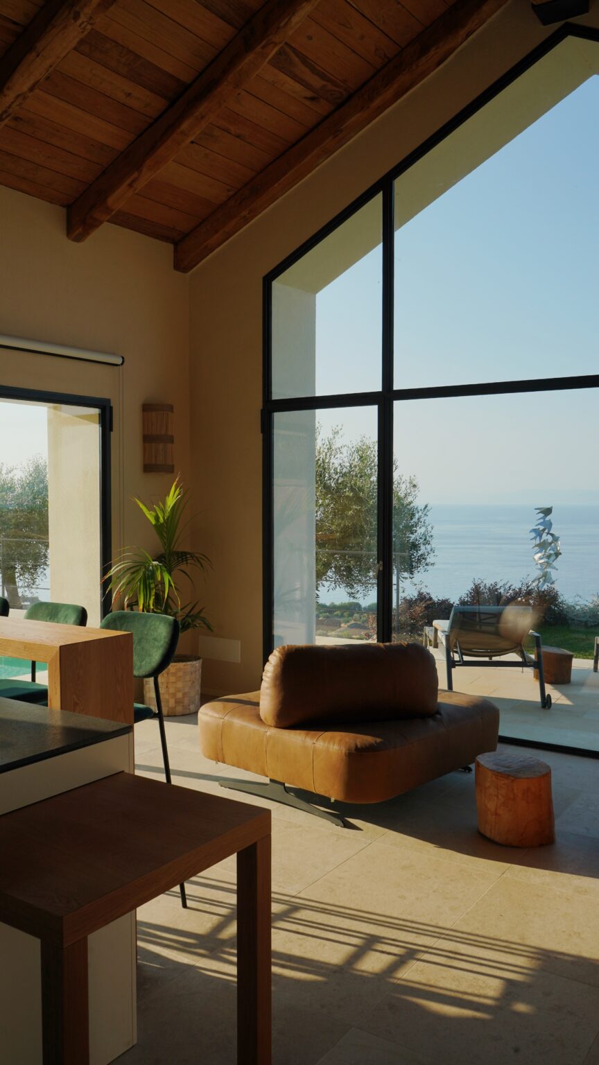 Modern living room with large windows showcasing a serene ocean view, featuring a leather lounge chair, wooden dining table, and indoor plants, illuminated by natural light. | Only on SopranoVillas