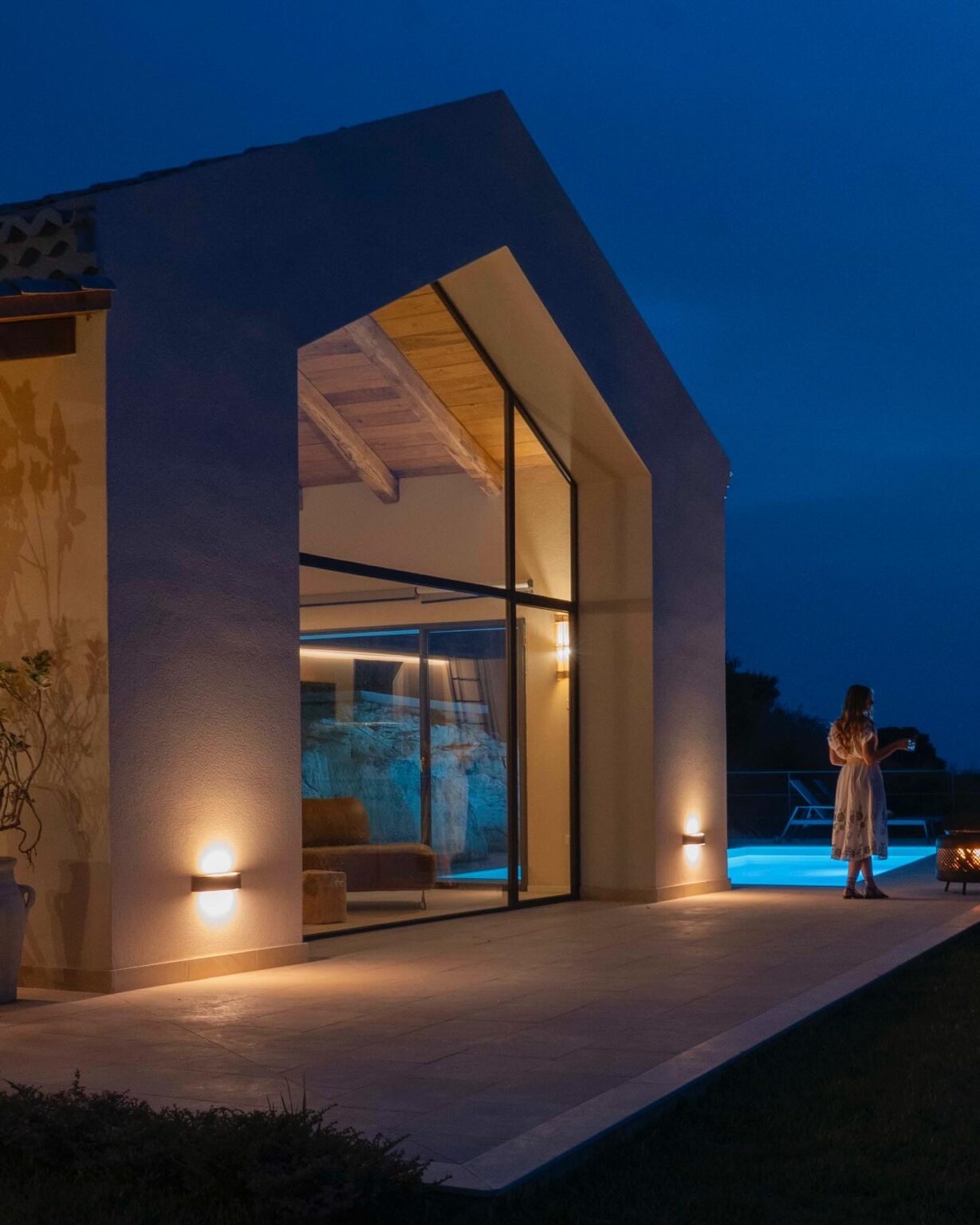 Modern villa exterior at night with large windows, illuminated by outdoor lights, and a woman walking towards a swimming pool. | Only on SopranoVillas