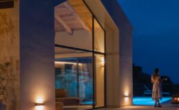 Modern villa exterior at night with large windows, illuminated by outdoor lights, and a woman walking towards a swimming pool.