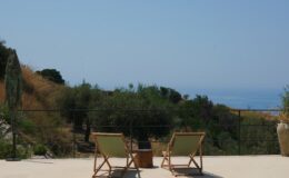 Two lounge chairs on a terrace overlooking a serene landscape with trees and ocean in the background, perfect for relaxation and enjoying the view.
