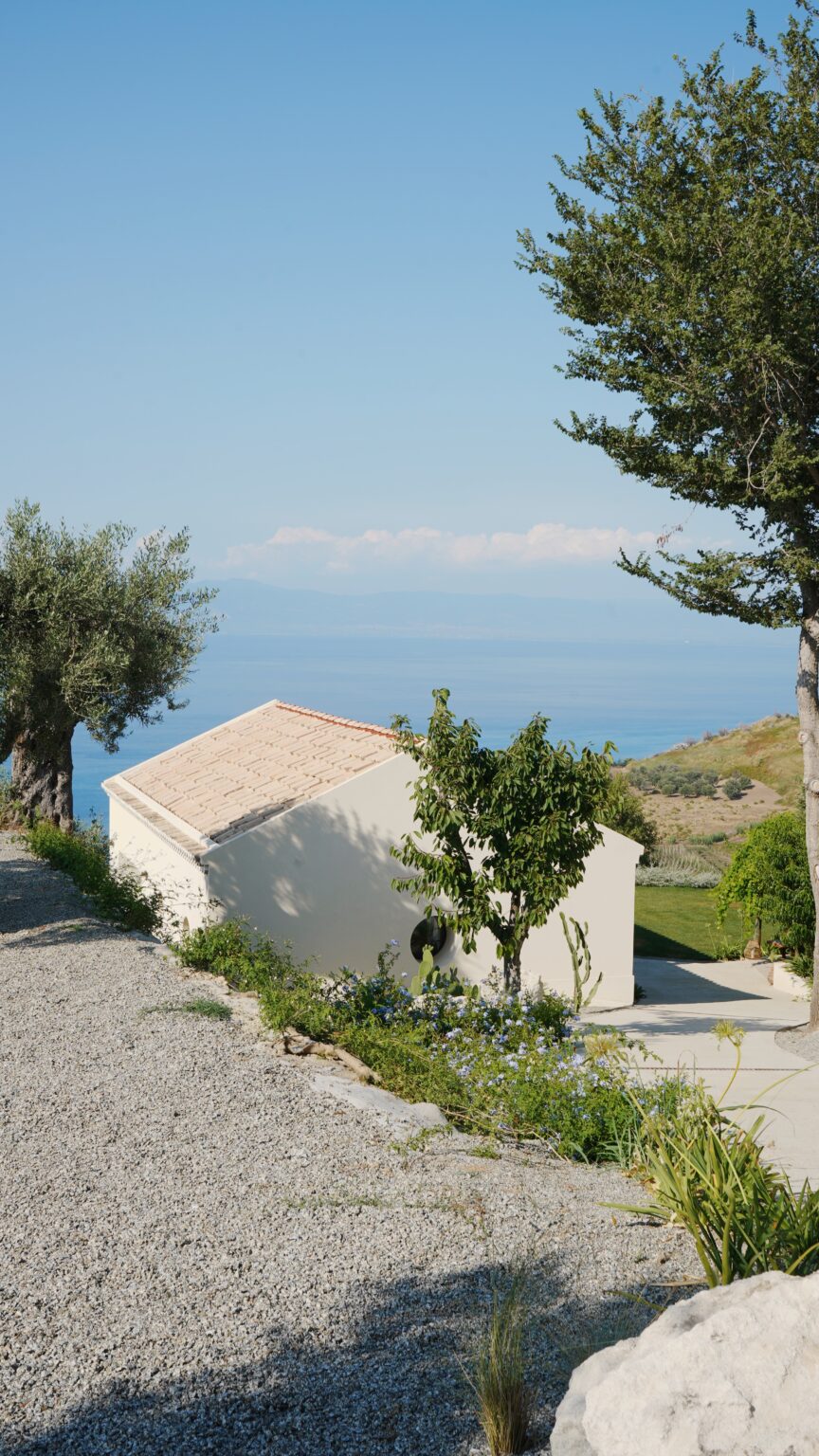 Scenic view of a white house on a hillside, surrounded by greenery and olive trees, overlooking a tranquil blue sea and distant mountains under a clear sky. | Only on SopranoVillas
