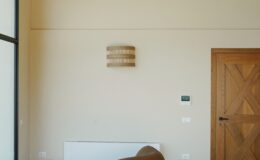 Interior view of a modern living space featuring a textured wall sconce, wooden door, and minimalistic heating unit, highlighting a neutral color palette and contemporary design elements.