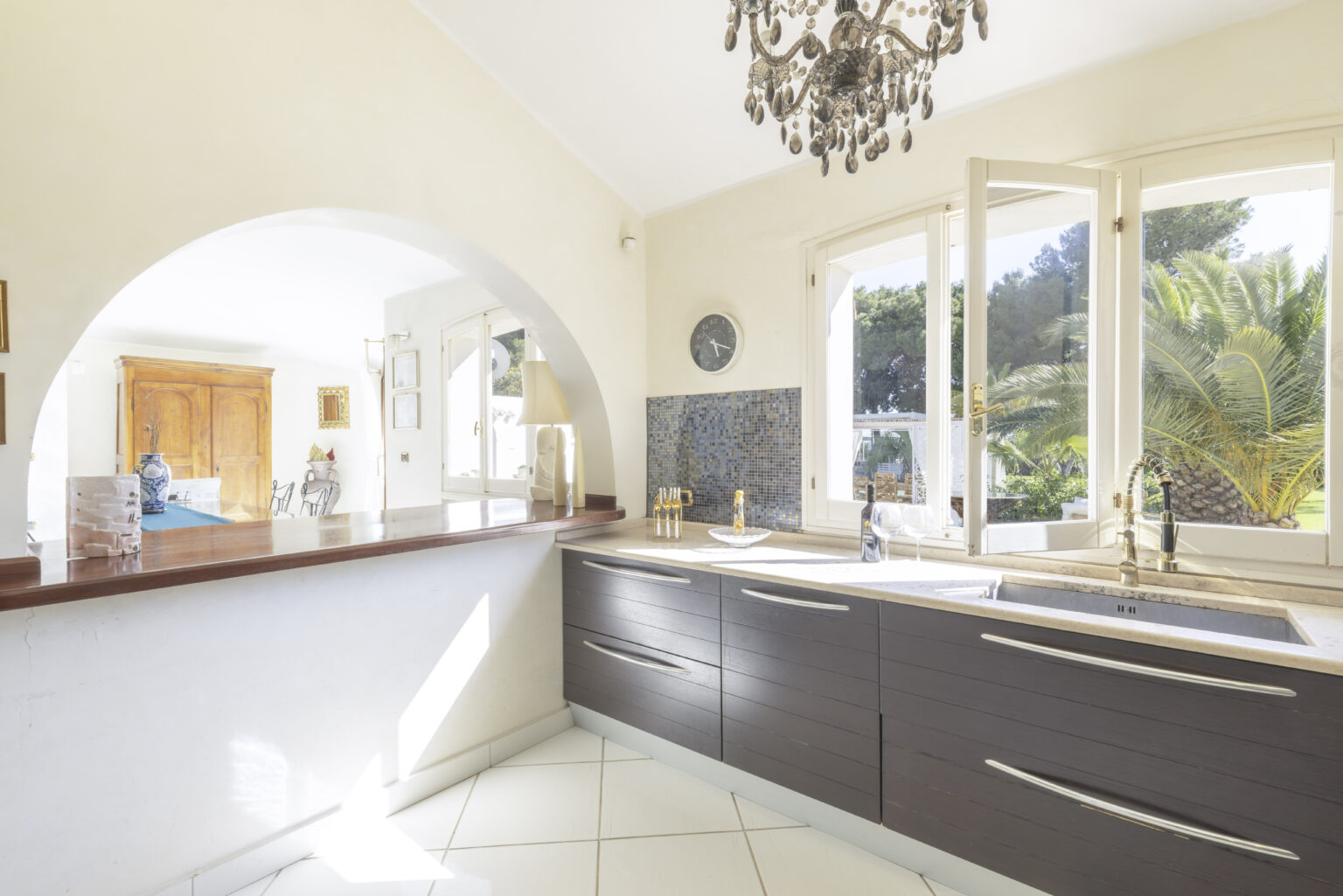 Modern kitchen interior featuring dark wood cabinets, stainless steel fixtures, and large windows that provide natural light and a view of greenery. The space includes a breakfast bar and elegant chandelier, highlighting a stylish and functional design. | Only on SopranoVillas