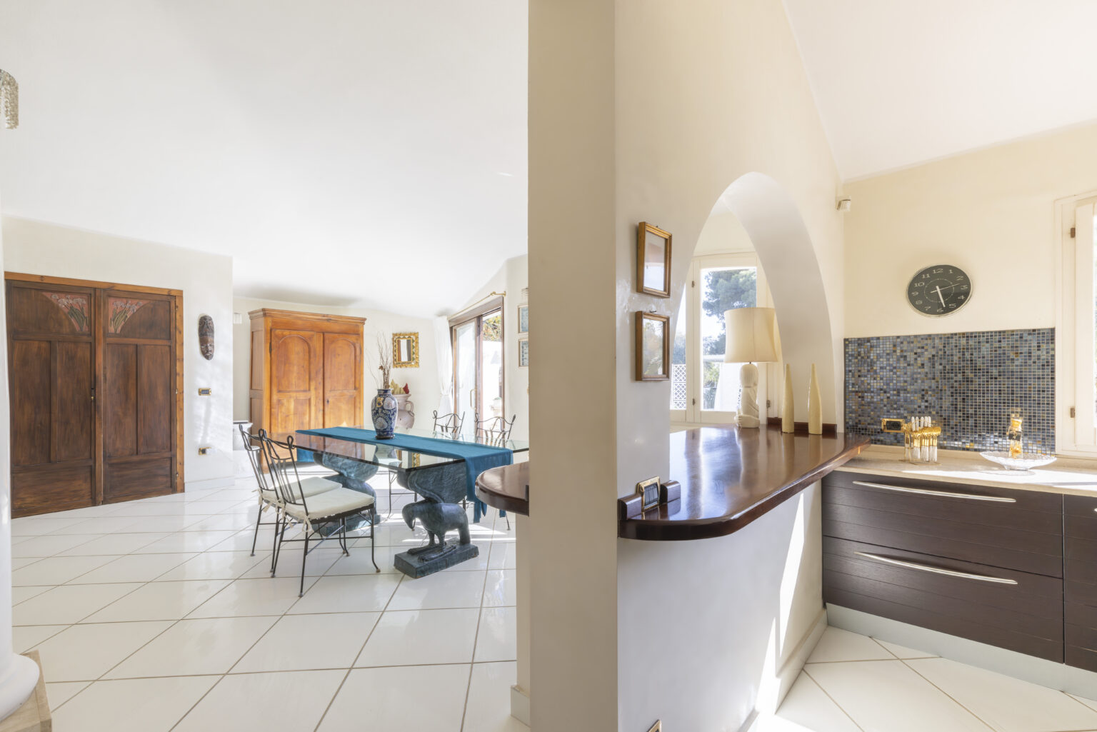 Modern open-concept living space featuring a dining area with a blue table and elegant chairs, adjacent to a stylish kitchen with dark cabinetry and a breakfast bar, illuminated by natural light. | Only on SopranoVillas