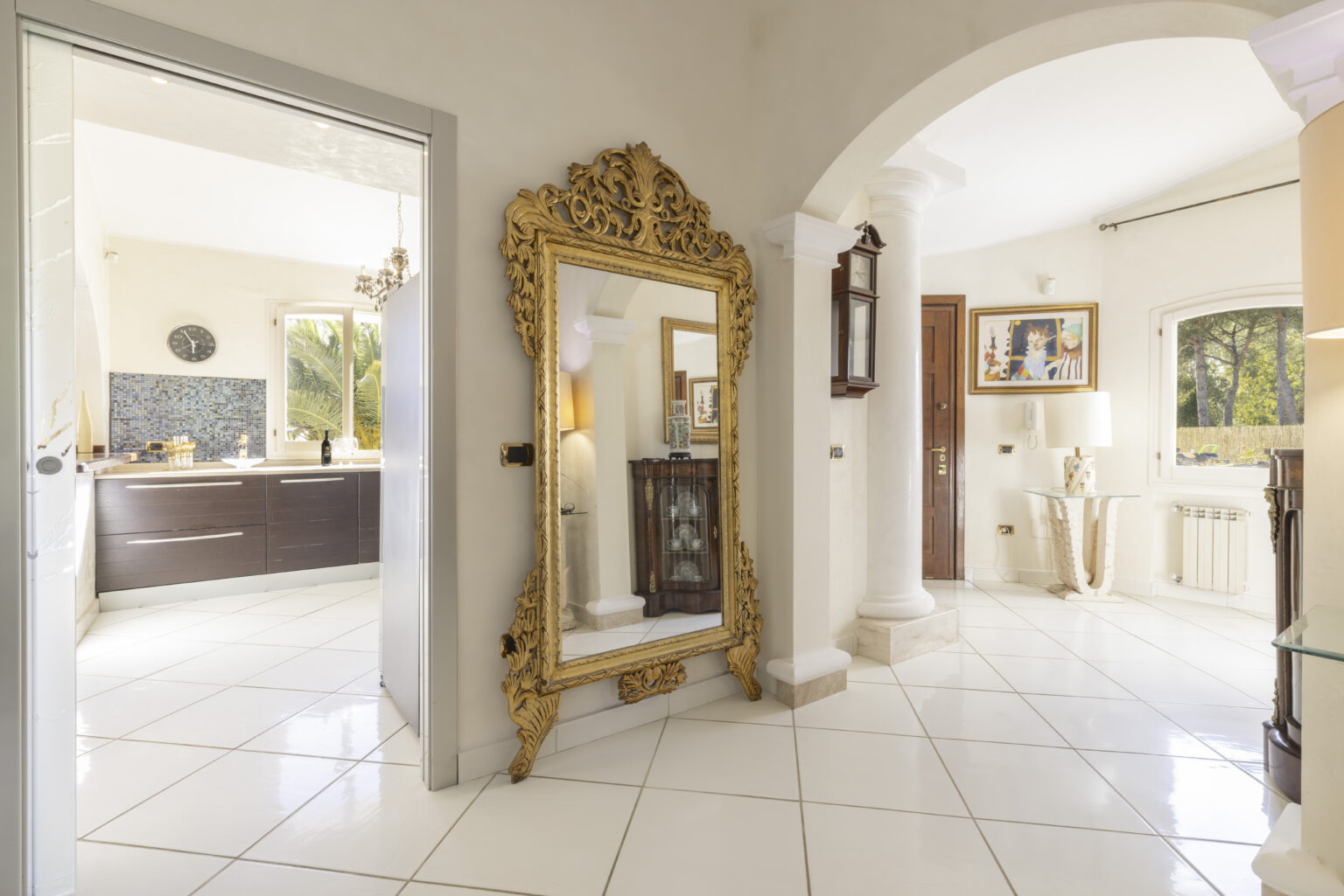 Elegant entryway featuring a large ornate gold mirror, leading to a modern kitchen area with dark cabinetry. The bright, open space showcases white tiled flooring and decorative art pieces, creating a welcoming atmosphere. | Only on SopranoVillas