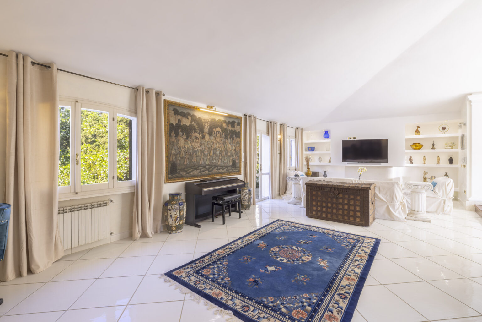 Bright and spacious living room featuring large windows, elegant curtains, a grand piano, and a decorative rug. The room is adorned with modern decor and a mounted flat-screen TV, creating a welcoming atmosphere. | Only on SopranoVillas