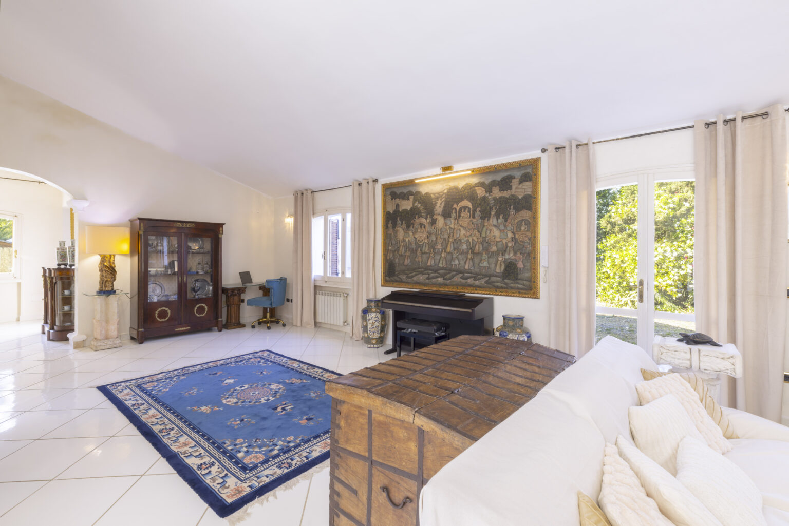 Spacious living room featuring a vintage wooden coffee table, a large blue area rug, elegant curtains, and a grand piano, with natural light streaming through large windows and a decorative wall tapestry. | Only on SopranoVillas