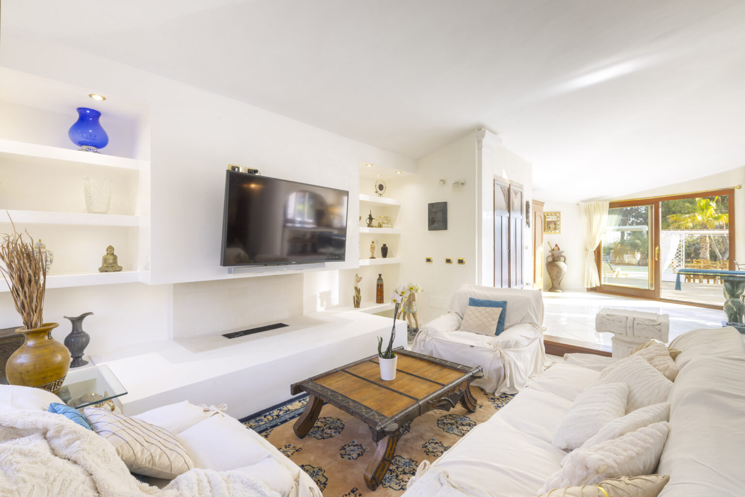 Bright and airy living room featuring white sofas, a wooden coffee table, and a modern TV unit. Natural light floods the space through large windows, complemented by decorative shelves and a cozy rug. Ideal for relaxation and entertainment. | Only on SopranoVillas