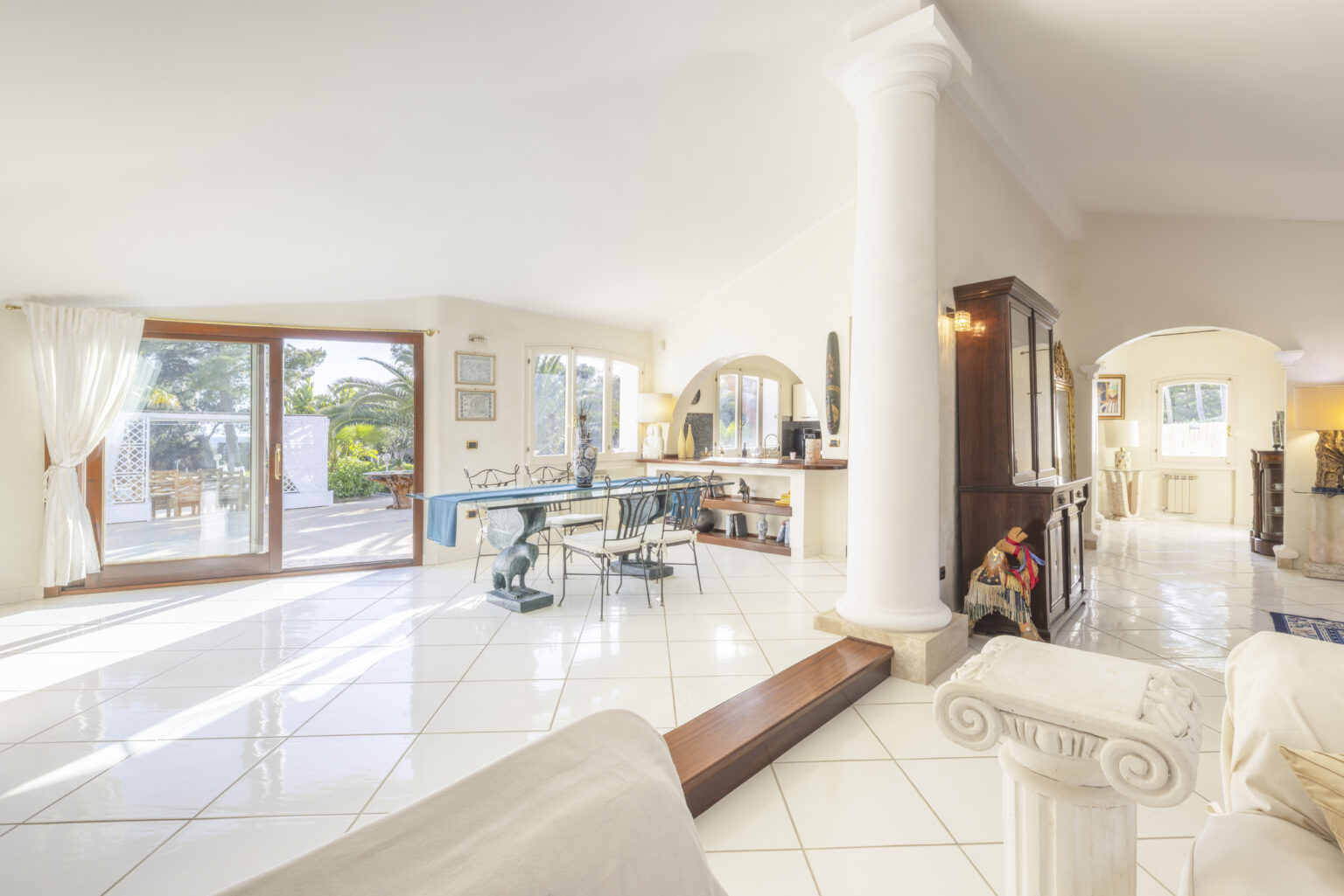 Spacious modern living area featuring large windows, white tiled floors, and elegant decor. The room includes a dining table with metal chairs, a stylish bar area, and a view of an outdoor patio surrounded by greenery. Natural light enhances the inviting atmosphere. | Only on SopranoVillas