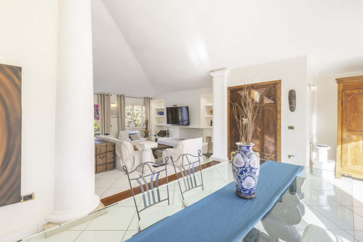 Bright and airy interior of a modern living space, featuring a glass dining table with a blue table runner and decorative vase, open to a cozy lounge area with a television and stylish furnishings. Natural light floods the room through large windows, highlighting the contemporary design and warm color palette. | Only on SopranoVillas