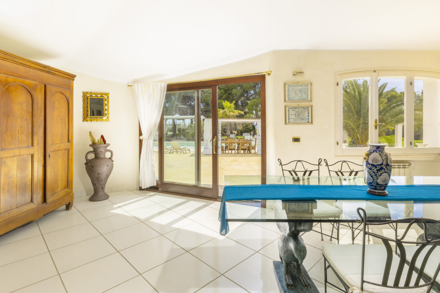 Bright and airy dining area featuring a glass table with a blue table runner, elegant metal chairs, and a decorative blue vase. A wooden cabinet and an ornate mirror add charm, while large windows provide a view of the outdoor patio and greenery. Ideal for showcasing modern interior design with a touch of classic style. | Only on SopranoVillas