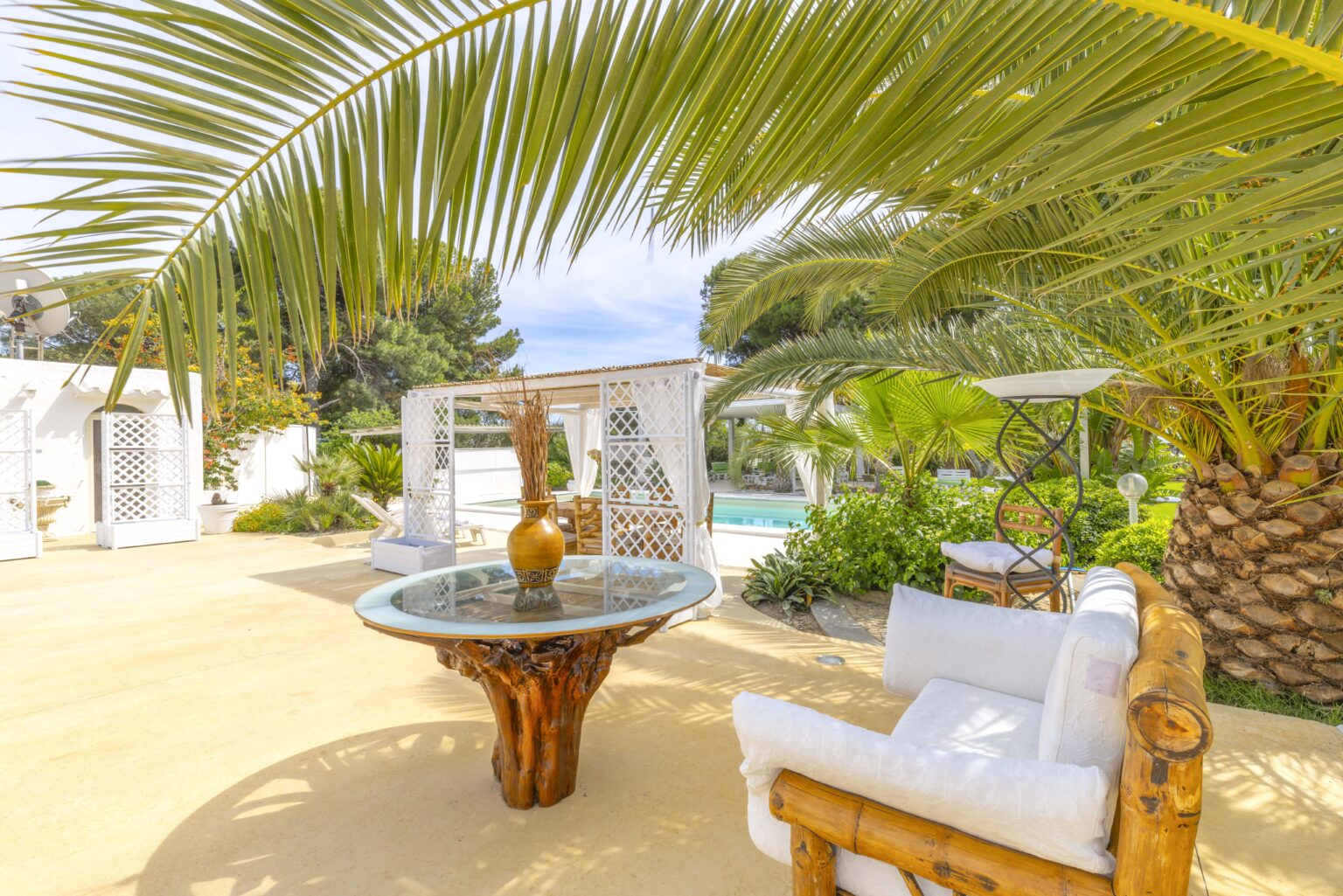 Outdoor patio featuring a wooden table and comfortable seating surrounded by lush palm trees and a swimming pool in the background, ideal for relaxation and leisure. | Only on SopranoVillas