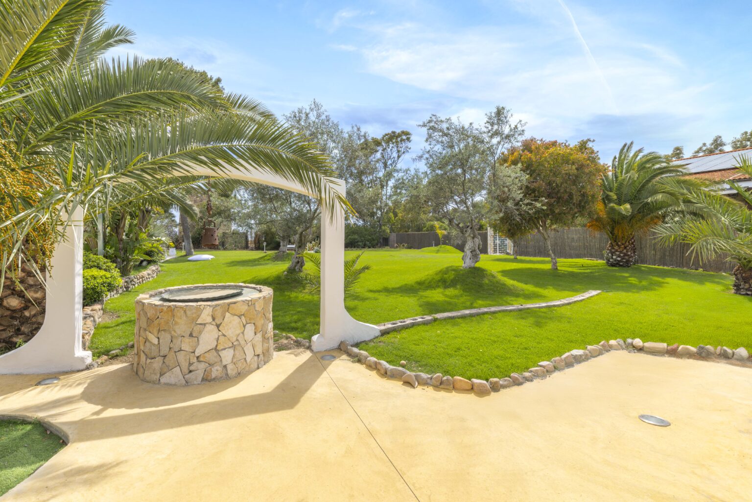 Lush green backyard with palm trees, a stone well, and a spacious lawn under a clear blue sky, ideal for relaxation and outdoor activities. | Only on SopranoVillas