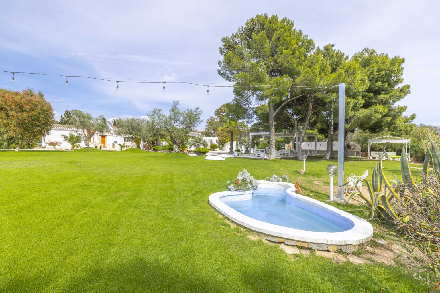 Lush green garden featuring a small outdoor jacuzzi surrounded by trees and decorative lighting, ideal for relaxation and outdoor gatherings. | Only on SopranoVillas