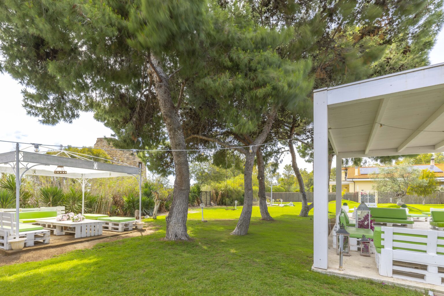 Lush green garden featuring tall pine trees, a spacious lawn, and a modern outdoor seating area with a pergola in the background. Ideal for relaxation and outdoor gatherings. | Only on SopranoVillas